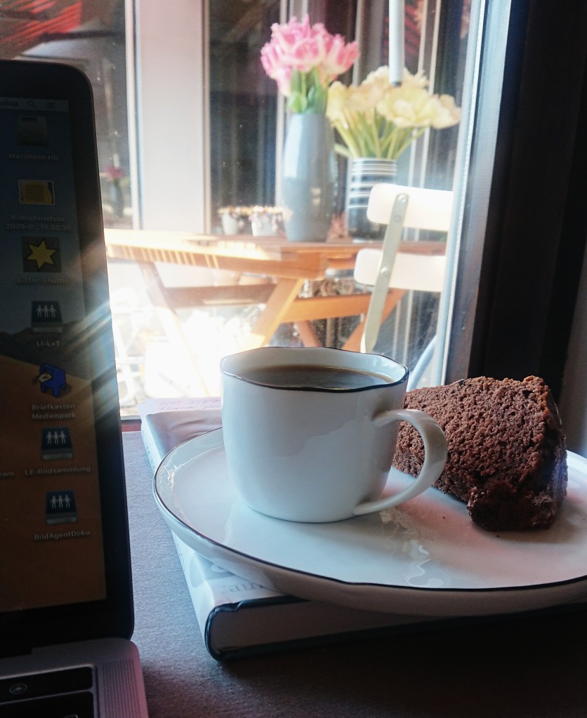 Ein Stück Gugelhupf auf einem weißen Teller. Dazu eine Tasse Kaffee und ein Blick nach draußen auf eine Terrasse. 